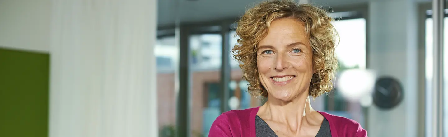 Eine Frau steht mit verschränkten Armen in einem Büro und schaut lächelnd in die Kamera.