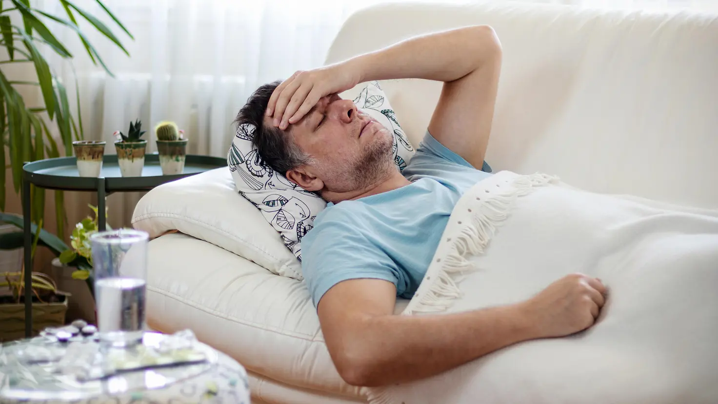 Mann liegt mit geschlossenen Augen und sichtlich erschöpft auf Sofa und hält sich die Hand an die Stirn.